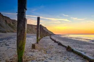 A beach at sunset features wooden posts extending into the distance, with cliffs on the left and a calm sea on the right – an idyllic setting perfect for anyone in the wedding planning business looking to create unforgettable moments.
