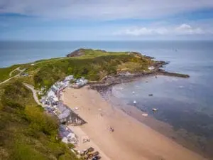 Aerial view of a coastal village with sandy beach, small white buildings, scattered boats on the shoreline, and green hills extending into the ocean.