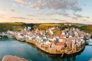 A coastal village with red-roofed houses is situated along a curved shoreline bordered by calm water and green hills under a partly cloudy sky.