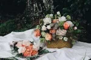 A wicker basket filled with assorted flowers including peach roses and white peonies sits on a white cloth next to a bouquet of similar flowers. Green foliage is visible in the background.