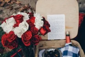 A bouquet of red and white roses sits next to a handwritten letter, a bottle of wine, and a picnic basket with a blue and white checkered cloth.