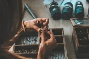 A person is examining a piece of jewelry with various other jewelry items laid out in a wooden organizer on the table. Rolls of thread and a tool are visible in the background.