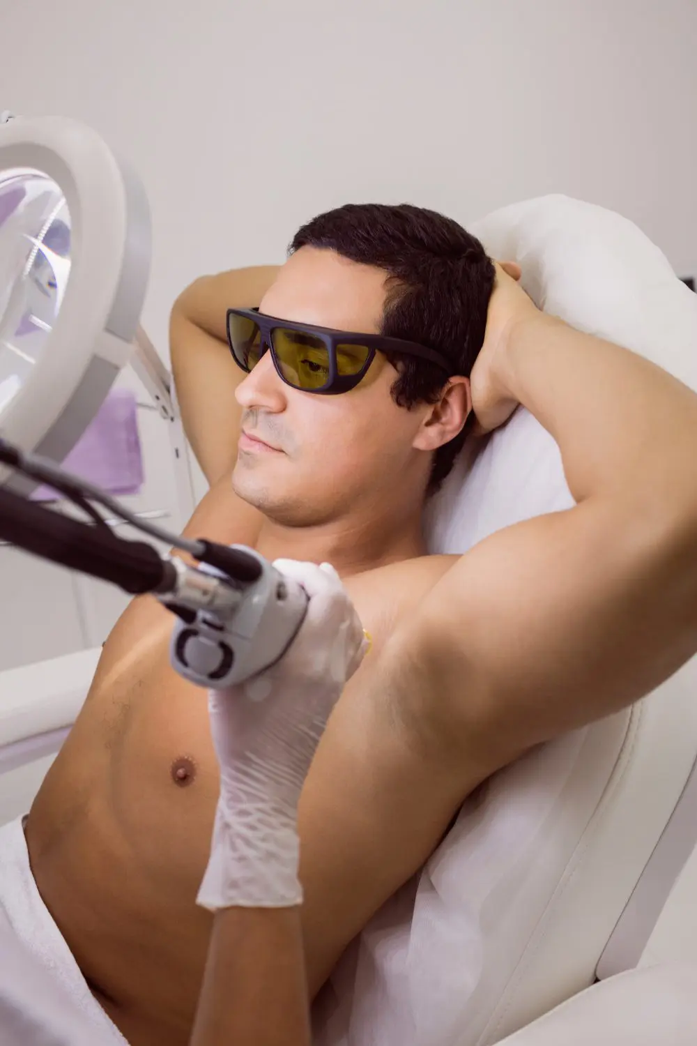 A man undergoing a laser hair removal treatment on his underarm, wearing protective eyewear, while lying on a white medical chair, with a clinician's gloved hand operating the laser device.
