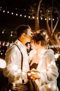 A man in a white shirt and black suspenders kisses a woman's forehead. She wears a white fur coat. They are surrounded by string lights at night.