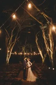A bride and groom stand under illuminated string lights and tall trees at an outdoor wedding ceremony at night.