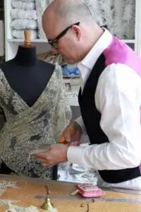 A man is working with a dress on a mannequin. He wears a white shirt, black vest, and glasses, and holds scissors in his hand. Fabric bolts and sewing materials are visible in the background.