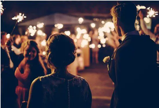 Bride and groom stand before a crowd holding sparklers during an evening outdoor event.
