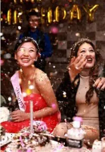 Two women smile and celebrate amid a flurry of confetti at a birthday party. A "Happy Birthday" banner is visible in the background.
