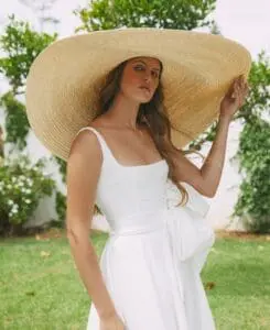 A woman in a white dress and large straw hat stands outdoors in a garden, looking directly at the camera. There are green plants and a white wall in the background.