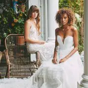 Two women in white gowns are seated on wicker furniture in an elegant, greenery-filled setting.