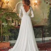 A bride stands with her back to the camera, wearing a long white gown with a deep V-back and lace sleeves. She is in a room adorned with plants and vintage furniture.