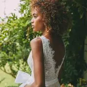 A woman with curly hair stands outdoors in a white lace dress with a low back, looking to the side. Green bushes and foliage are in the background.