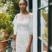 A woman in a white lace dress stands outside near a building, surrounded by greenery and potted plants.