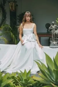A woman in a white dress with a bow sits on a white wall surrounded by greenery and a sculpture.
