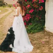 A woman in a white dress stands on a dirt path with her back to the camera, near pink flowers and greenery, while a black and white dog sits beside her.