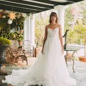 Woman in a white wedding dress standing under a porch with several chairs, plants, and decorative items in the background.