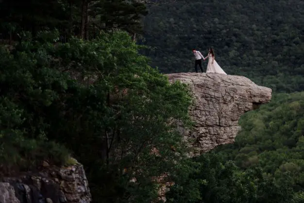 A couple stands on a rocky cliff edge, surrounded by lush green trees and a dense forest in the background. The woman wears a white dress and the man wears a white shirt and dark pants.