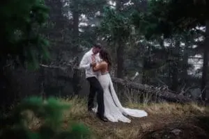 A bride and groom, dressed in wedding attire, share an intimate moment in a misty forest setting. The bride wears a white gown, and the groom is in a white jacket and dark pants. Green trees surround them.