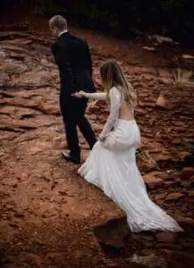A couple dressed formally, a man in a black suit and a woman in a white dress, walking on a rocky terrain. The woman holds the man's hand as they proceed.