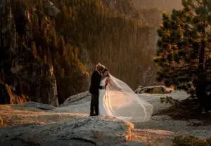 A bride and groom stand close together on a rocky cliff at sunset, surrounded by trees and overlooking a forested valley. The bride's veil is flowing in the wind.