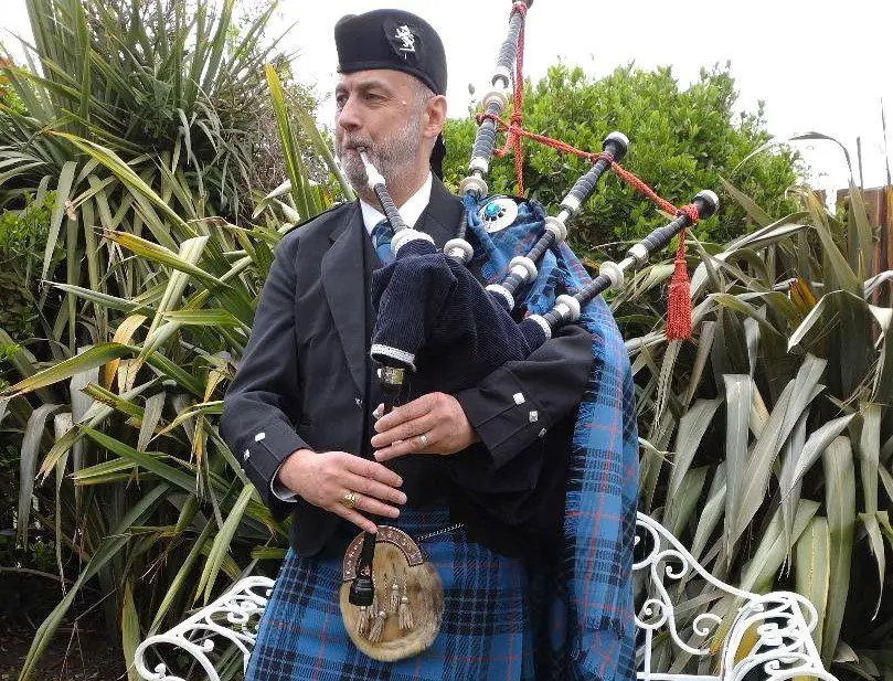 A man wearing traditional Scottish attire is playing a bagpipe, providing enchanting melodies for a wedding ceremony set amidst tall plants.