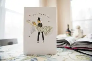A greeting card with an illustration of a bride and groom dancing, accompanied by the text "Celebrating With You!" rests on a table next to an open book and a bottle.