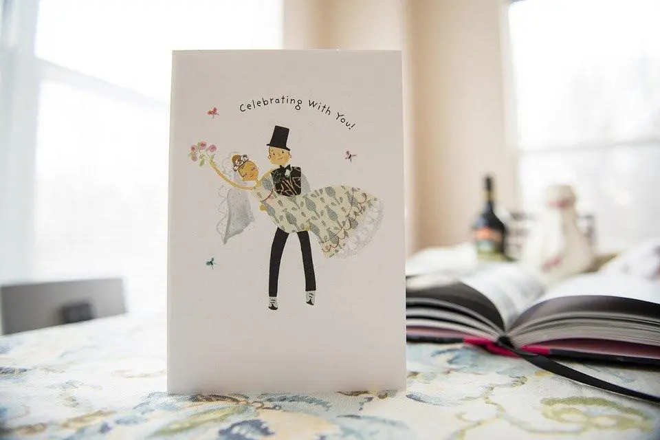 A greeting card with an illustration of a bride and groom dancing, accompanied by the text "Celebrating With You!" rests on a table next to an open book and a bottle.