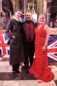 Three people pose indoors in front of a British flag. One wears a long coat, one is in a suit, and the other is in a red dress.