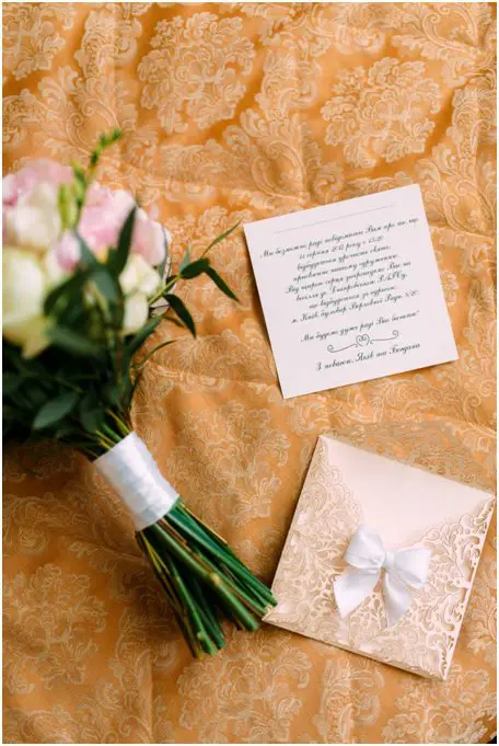 A bouquet of flowers, a wedding invitation card, and an envelope with a white bow are placed on an ornate, gold-patterned fabric background.