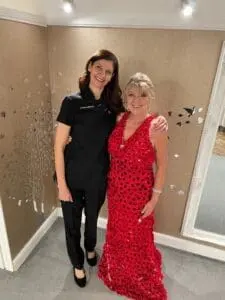Two women stand side by side; one is wearing a black outfit and the other is in a red, patterned dress. They are indoors with a wall adorned with small decorations in the background.
