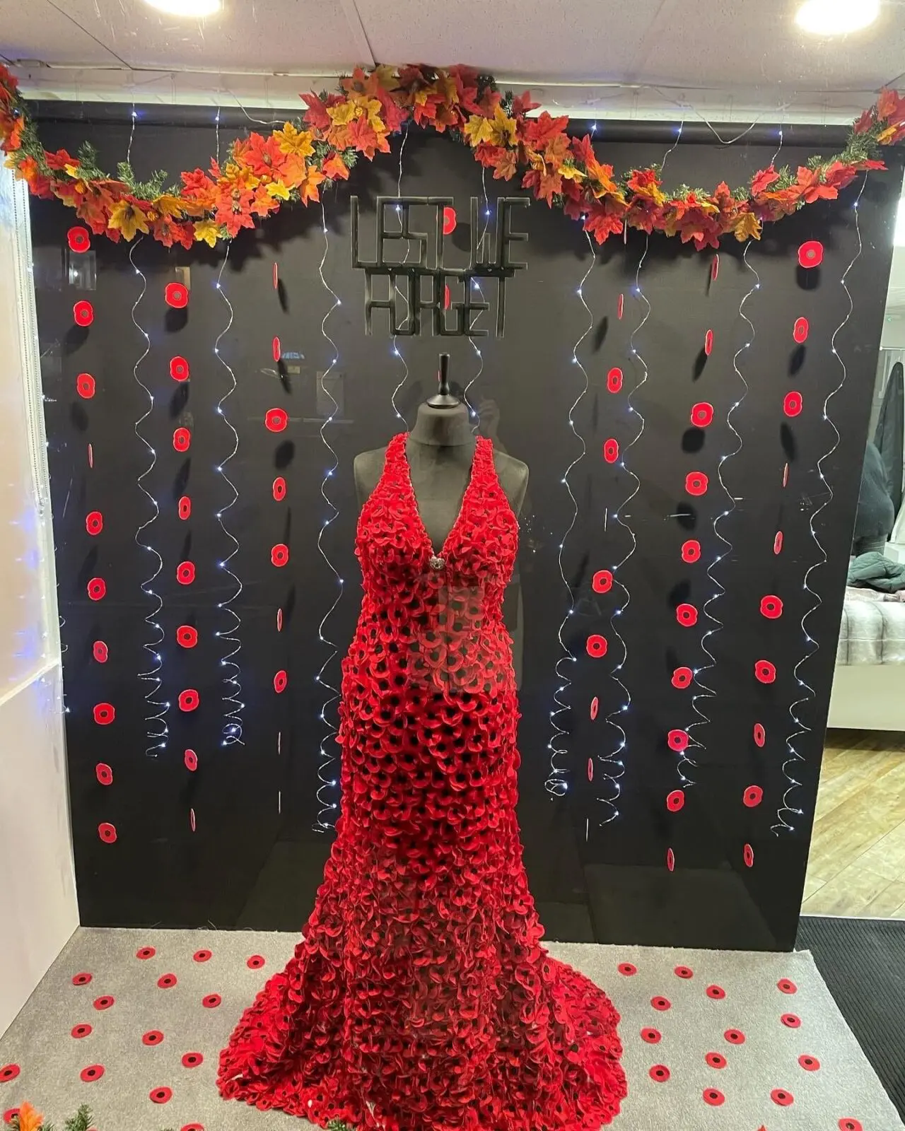 A red dress covered in a pattern of red flowers is displayed on a black mannequin. It's set against a black and floral-themed backdrop with leaf garlands and string lights.