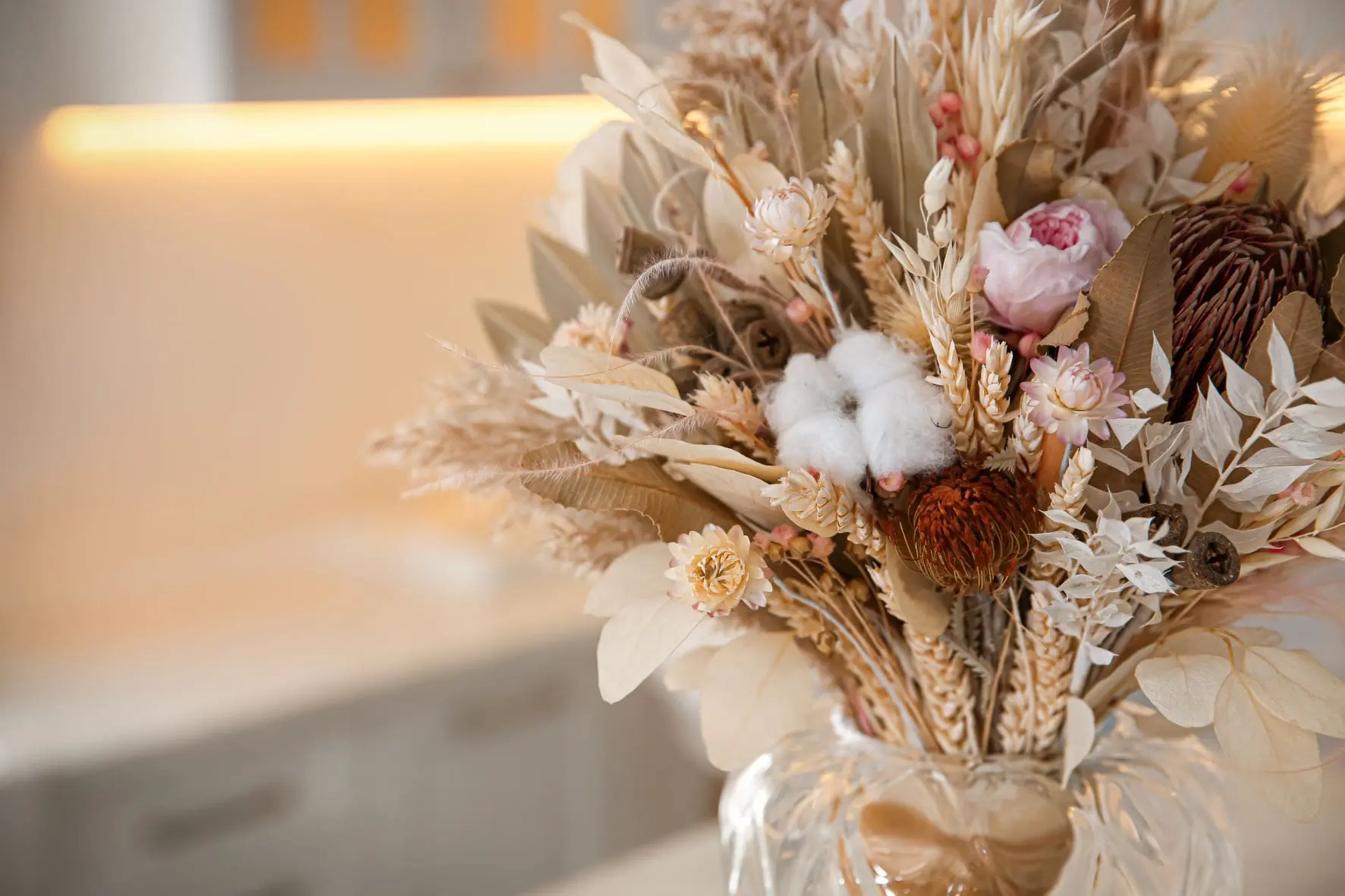 A bouquet of dried flowers, featuring various beige and pink blossoms and cotton plants, arranged in a clear glass vase.
