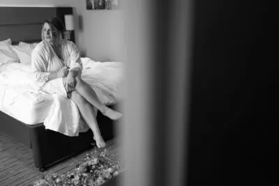 A person sits on the edge of a bed in a hotel room, wrapped in a robe, looking out of the frame. The room appears tidy, and there is a bouquet of flowers in the foreground. The image is in black and white.