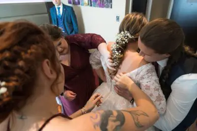 A bride getting ready with the help of three women; one adjusting her gown and the others attending to details, while a man in a blue suit stands in the background.