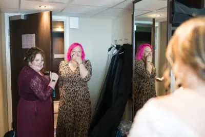 Two women are in a room, one is wearing a leopard print dress and has pink hair, the other is wearing a maroon dress. They are looking at someone off-camera. Clothing is hanging in the background.