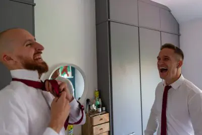 Two men in dress shirts and ties are laughing together in a room with grey wardrobes and a small dresser.