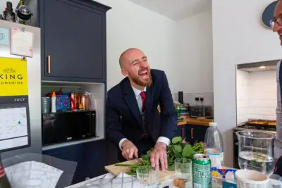 Man in a suit laughing while chopping vegetables in a modern kitchen with dark cabinets and a calendar on the fridge.