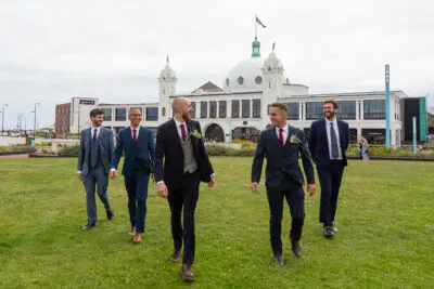 Five men in suits walk across a grassy area toward a historic white building with domes and a flagpole on top.