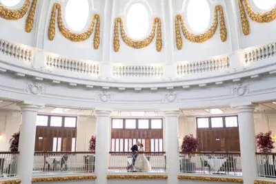 A couple shares a moment on a balcony inside an ornate, white domed room with large windows and gold accents.