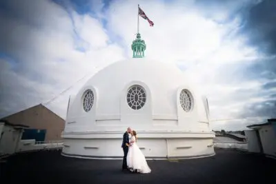A couple dressed in wedding attire stands on a rooftop in front of a large white dome with circular windows and a flag on top, under a partly cloudy sky.