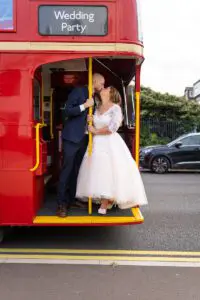 A couple, dressed in wedding attire, share a kiss on the rear platform of a red double-decker bus labeled "Wedding Party." The bride wears a white dress, and the groom is in a blue suit.