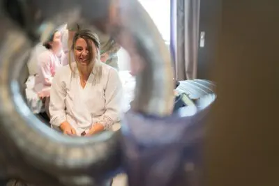 A woman in a white robe smiles and sits in a room, with a blurred foreground element creating a circular frame around her. Another person is visible in the background.