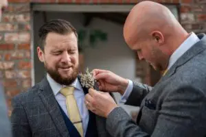 A man in a gray suit adjusts a boutonniere on another man in a gray suit with a yellow tie, who has an uncomfortable expression. Both are standing in front of a brick wall.