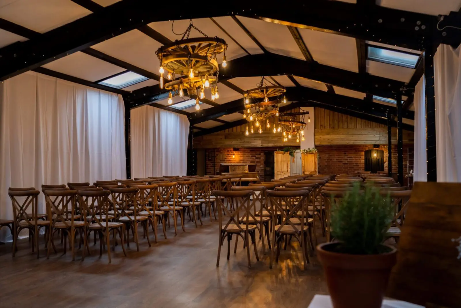 A rustic event space with wooden rafters, chandelier lighting, and rows of wooden chairs facing a focal point at the front. White drapes line the walls, and a potted plant is visible in the foreground.