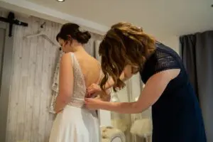 A woman helps another woman button the back of a dress in a room with light-colored walls.