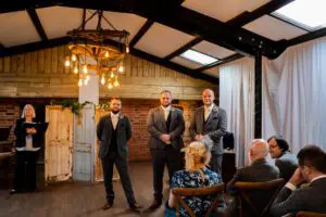 Indoor wedding ceremony scene with three men in suits standing at the front. Guests are seated facing them, and a person holding a book is to the left. The venue has exposed beams and warm lighting.