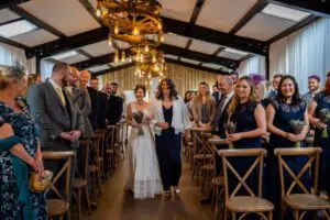 A bride walks down the aisle with an escort in a warmly lit, rustic venue lined with guests seated on wooden chairs.