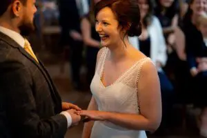 A bride and groom exchange rings during their wedding ceremony, with the bride smiling and guests blurred in the background.