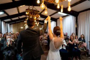 A couple raises their hands in celebration while facing seated guests in a well-lit hall decorated with hanging lightbulbs.