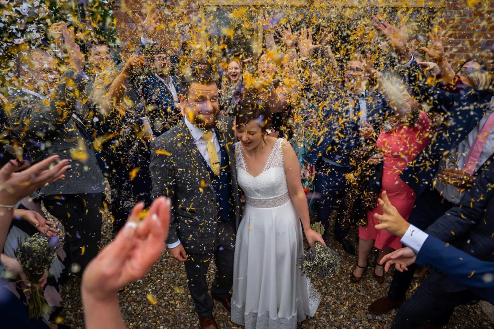 A newlywed couple is showered with confetti by a group of people. The bride is wearing a white dress, and the groom is in a suit. Both appear to be smiling and enjoying the celebration.